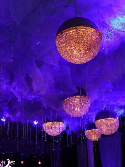A dramatic ceiling installation featuring spherical chandeliers suspended beneath a cloud of purple-lit fabric. This creative lighting solution adds depth and a unique visual focus to the event space, perfect for a modern and stylish reception.