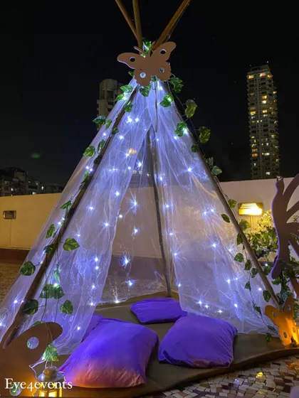 A close up of a whimsical teepee tent I created for a teen's butterfly themed birthday party. The tent is made of sheer fabric, illuminated with fairy lights, and decorated with green vines and a wooden butterfly cutout at the top.