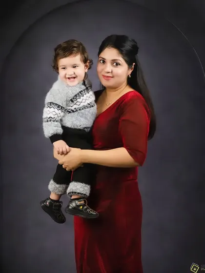 A mother holds her smiling son in her arms. Her elegant red dress complements the dark background, creating a rich and warm portrait.
