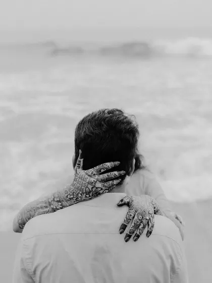 A powerful monochrome image focusing on the bride's intricate henna-covered hands as she embraces her groom from behind.