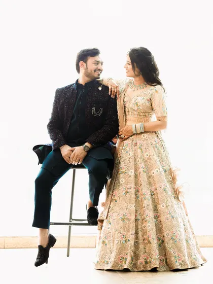 A bright and cheerful studio portrait against a clean white background. The playful interaction between the seated groom and standing bride creates a fun and relaxed pre-wedding photo.