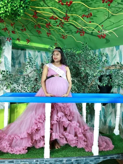 A serene maternity portrait. The mother-to-be, wearing a "Mom to be" sash and a lavender gown, sits peacefully on the bridge of our indoor garden set.