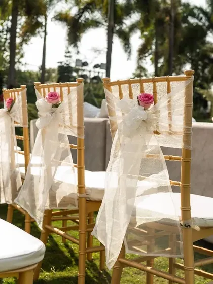 Elegant gold chiavari chairs decorated with delicate white organza sashes and a single pink rose for the wedding ceremony.