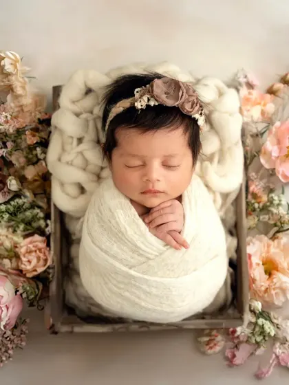 This newborn girl is wrapped snugly in a white swaddle, resting in a rustic wooden box surrounded by soft pastel flowers.