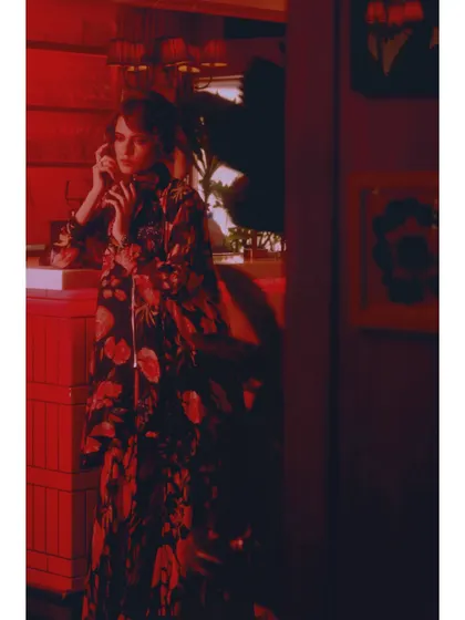 A vertical composition from 'The Wait', emphasizing the length of the floral coat and the atmospheric, shadow-filled room. Every element is styled to contribute to the narrative of waiting.