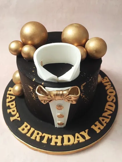 We present the essence of formal sophistication with our black and gold tuxedo cake. This design features a sharp, edible shirt collar and a lustrous gold bow tie set against a matte black tier, accented with gold splatters and spheres, making it an impeccable choice for a gentleman's milestone birthday.