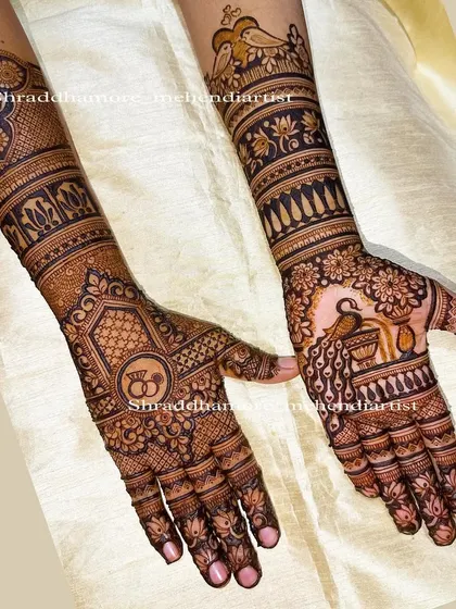 A beautiful engagement mehendi design featuring intertwined rings on one palm and a peacock with a diya on the other. The arms are adorned with lovebirds and lotus bands, telling a complete story.