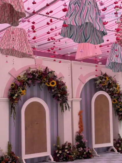 A whimsical stage for a Mayra ceremony, featuring classical English arches, a checkered floor, and an eclectic mix of patterned lampshades hanging from a pink ceiling.
