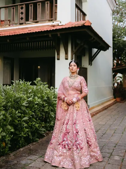 A full-length portrait of a bride in a beautiful pink floral lehenga, standing elegantly in front of a villa at the ITC Grand Goa.