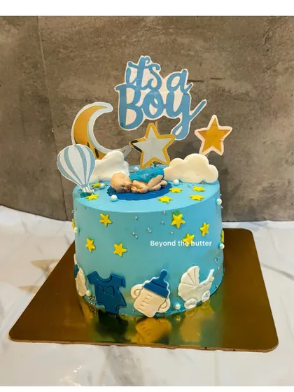 Welcome to the world, little one. This "It's a Boy" cake is decorated with a starry night theme, featuring a sleeping baby, fondant clouds, and cute baby-themed elements like a bottle and carriage.