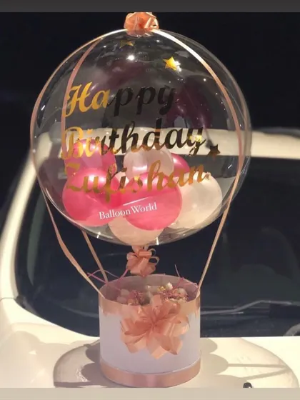 This hot air balloon hamper was designed for a birthday, featuring a personalized name in gold script on the clear balloon. The base is a simple, elegant box filled with flowers, perfect for evening delivery.