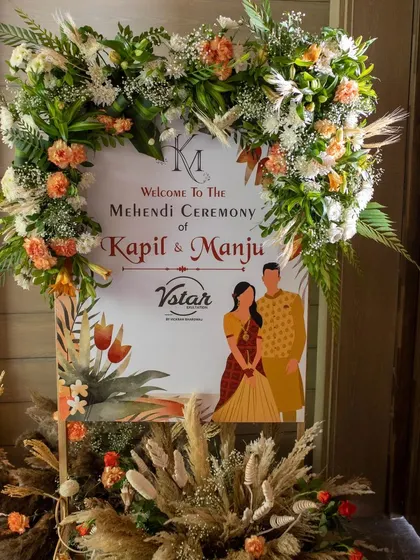 A beautiful welcome sign for a Mehendi ceremony, framed with a heart-shaped arch of rustic flowers and pampas grass.