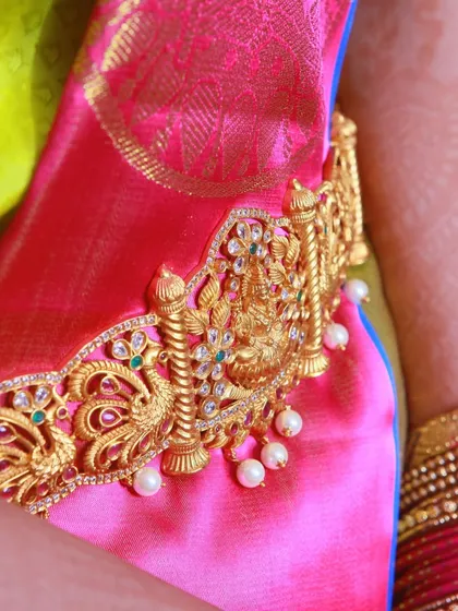 A close-up of a traditional gold Vaddanam (waist belt) with a goddess motif, worn over a bright pink silk saree.