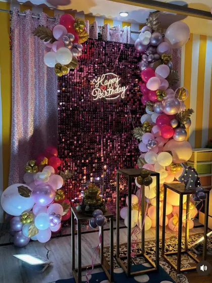 A stylish birthday setup with a pink shimmer wall and a modern balloon arrangement. The decor includes pedestals for placing the cake or other items.