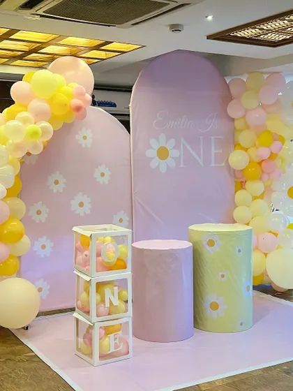 A sweet daisy-themed first birthday party. The decor features daisy-print backdrops, 'ONE' blocks filled with balloons, and a cheerful pink and yellow color palette.