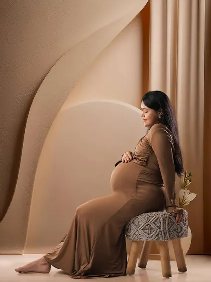 A graceful side profile in a simple brown gown. The abstract, neutral-toned background and modern stool create a contemporary and artistic maternity portrait.
