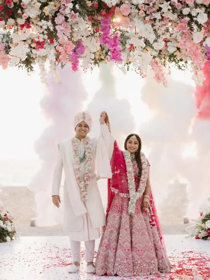 The couple stands triumphantly under a floral arch on the beach as pink and white smoke bombs go off behind them, creating a dramatic and celebratory image.