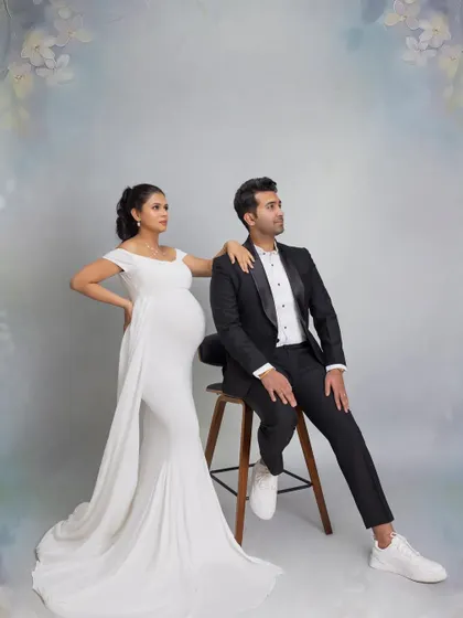 A chic and fashionable pose from a twin maternity shoot. The mother-to-be in a sleek white gown leans against her partner, who is seated, creating a stylish, magazine-worthy portrait.