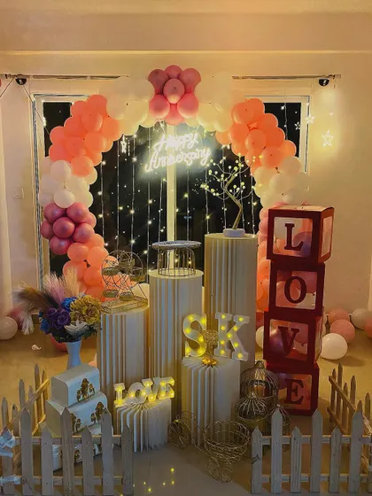 A soft and romantic anniversary decoration with a peach and white balloon arch. This setup includes 'LOVE' display boxes and a picket fence for a charming, rustic feel.