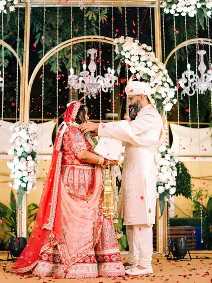 The garland exchange on a beautifully decorated stage. This wide shot captures the couple, the elegant floral and chandelier decor, and the significance of the Varmala ceremony.