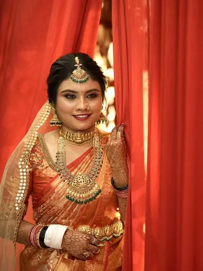 This bride is ready for her ceremony, peeking from behind a red curtain. She wears a rich orange silk saree with green-accented jewellery, and her makeup is bold and defined.
