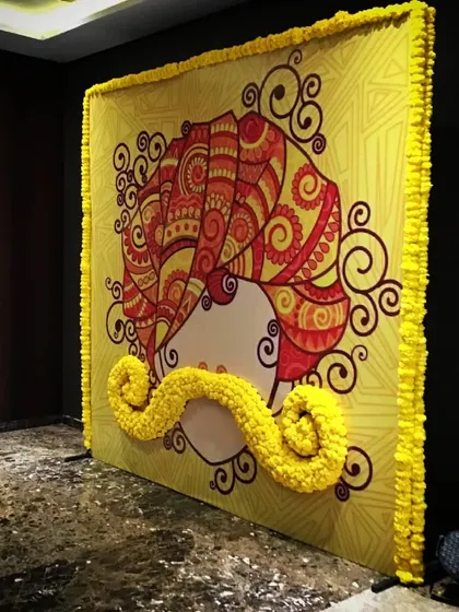A playful and artistic backdrop for a Mehendi, featuring a graphic of a man in a turban with a 3D mustache made from marigold flowers.