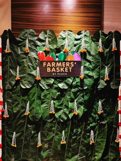 A "Farmers' Basket" photo wall made from a backdrop of large banana leaves and decorated with floral strings. This is a creative and on-theme backdrop for guest photos.