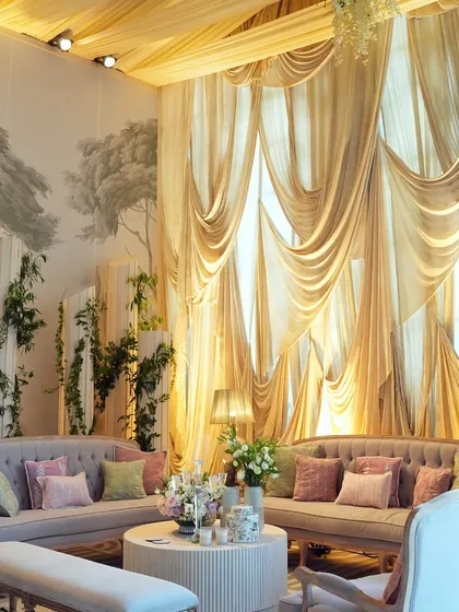 A luxurious lounge area set against a backdrop of hand-painted wallpaper and dramatic golden drapes. The pastel-colored sofas and floral arrangements create a soft, inviting space.