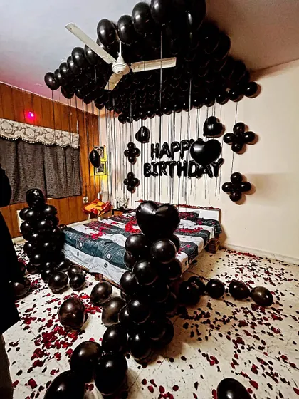 A dramatic all-black themed birthday room decoration. The setup includes black balloons covering the ceiling, balloon pillars, and rose petals on the floor.