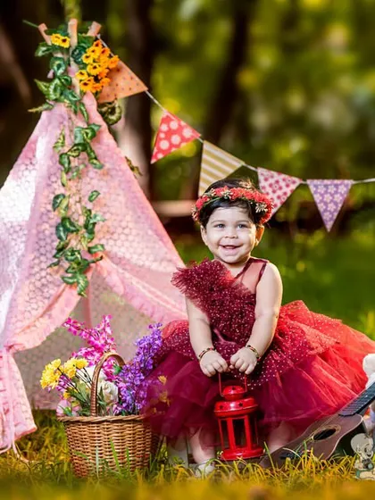 A toddler in a beautiful red dress sits in front of a pink teepee during her outdoor birthday photoshoot. The natural surroundings and festive props create a magical celebration.