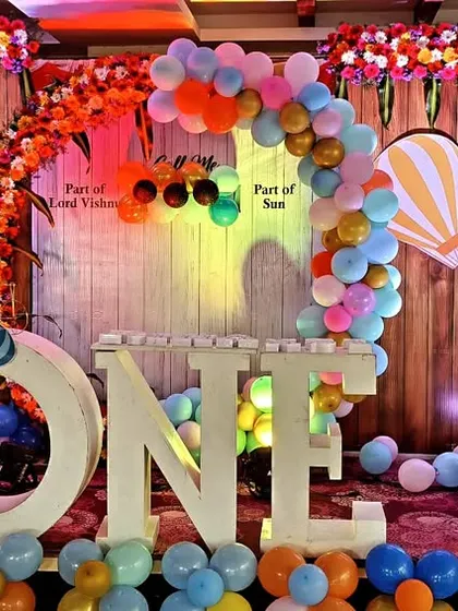 A vibrant and colorful first birthday party with a hot air balloon motif. The stage includes large "ONE" letters, elephant cutouts, and a multi-colored balloon arch, perfect for a lively celebration.