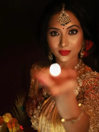 A beautiful and symbolic shot for Deepavali. The model's face is illuminated by the diya, highlighting her glowing makeup, bold red lip, and sparkling eyes. This is a perfect example of festive glamour.