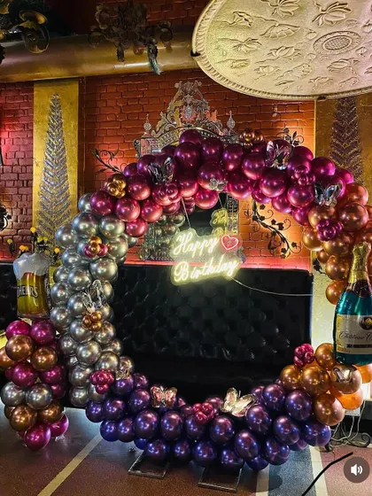 A lavish birthday party decoration in a lounge. The setup features a large circular balloon frame with a mix of metallic colors like purple, magenta, silver, and rose gold.