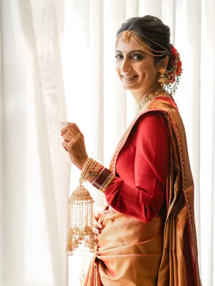 A bride in a beautiful red and gold saree, looking out the window with a gentle smile. This is a classic, elegant portrait filled with warmth.