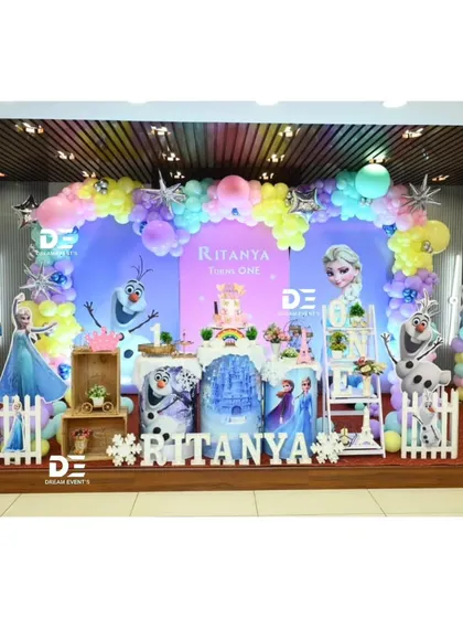A winter wonderland with this 'Frozen' theme for Ritanya's first birthday. The setup features Elsa, Anna, and Olaf, a snowy backdrop, and a beautiful balloon arch in icy shades of blue, purple, and white.