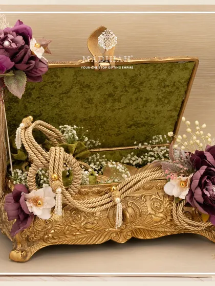 An ornate Rajwada chest in gold finish, decorated with purple flowers and elegant tassels. The olive green velvet interior adds a touch of classic sophistication to this wedding hamper.