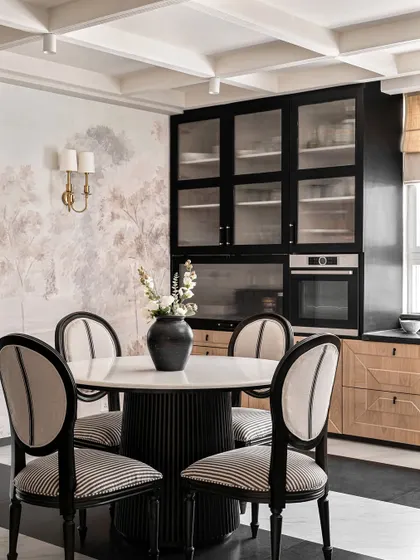The informal dining area in Riviera Maison, where we paired a fluted-base marble-top table with classic striped dining chairs. The black and oak cabinetry and a soft landscape mural create a sophisticated, European-inspired look.