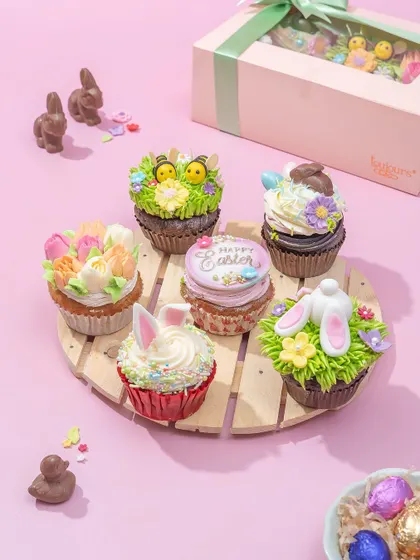 A set of six adorable Easter cupcakes on a wooden platter, decorated with buttercream grass, bunny ears, and tiny chicks. These are a whimsical and delicious addition to your dessert table.