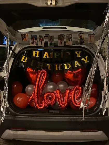 Surprise on the go! This is a birthday car trunk decoration, filled with red and silver balloons, a 'Happy Birthday' banner, a large 'Love' foil balloon, and personalized photos hanging from the top.