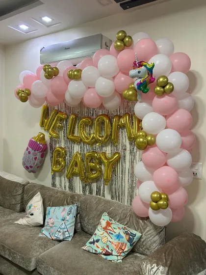A classic "Welcome Baby" decoration for a living room. This setup features a pink, white, and gold balloon arch over the sofa, a silver foil curtain backdrop, and large gold foil letters.