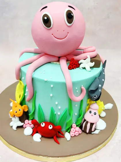 We create an ocean of fun with this adorable pink octopus cake. The cheerful octopus, with its big, friendly eyes, is the star of this underwater scene, surrounded by a crab, fish, and other sea creatures on a beautiful aqua-blue tier.