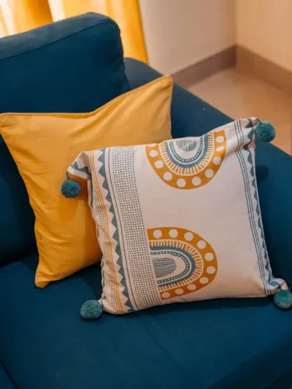 A close-up of our 'Aura' cushion cover with its playful pom-pom details, paired with a solid mustard cushion. This combination adds a pop of color and pattern to the bold teal sofa.