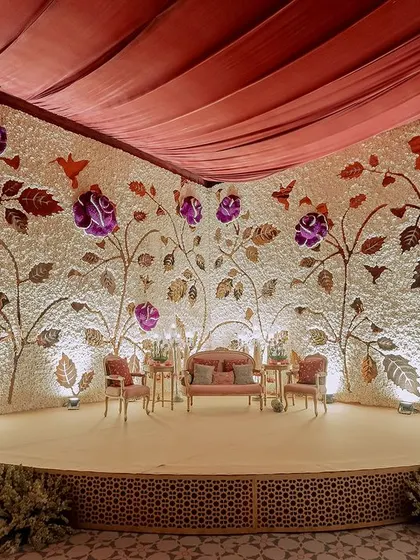 This magnificent stage backdrop is a custom-made flower wall, with a base of white carnations and a design of climbing roses made from purple and metallic-gold dyed petals.