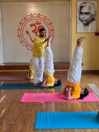 An instructor provides support and guidance as students hold Sarvangasana (Shoulderstand), another key inversion in the Hatha yoga sequence.