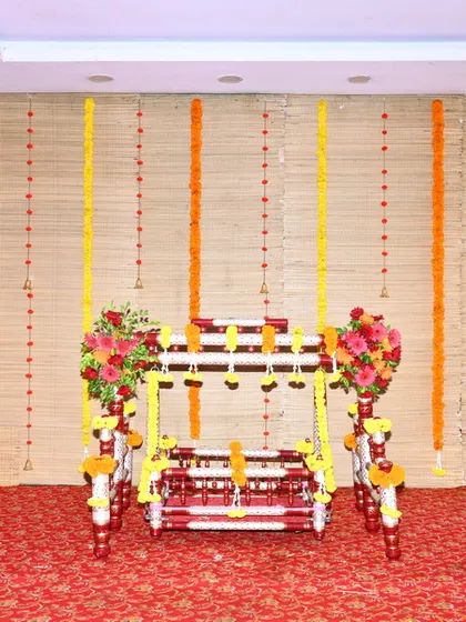 A simple and traditional wooden cradle for a naming ceremony, decorated with red and yellow flowers and set against a backdrop of marigold garlands. This is a standard rental option for indoor functions.
