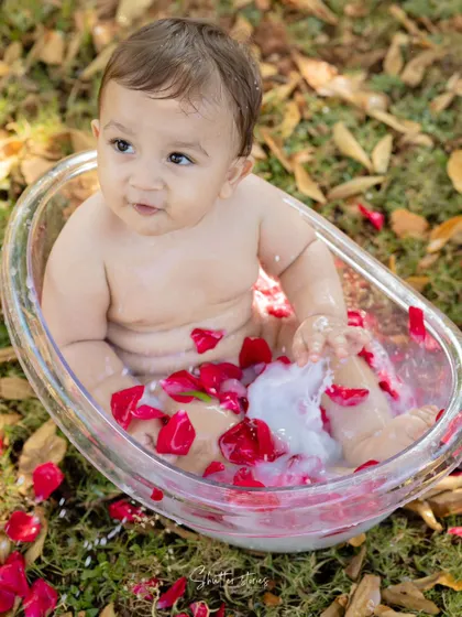 A baby boy enjoys a milk bath with red rose petals in a beautiful outdoor setting.