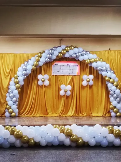A simple and elegant stage decoration for a school or community event. This setup features a large balloon arch in white, gold, and silver over a golden curtain backdrop.