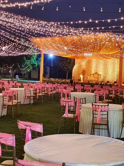 An expansive outdoor reception setup under a canopy of fairy lights. The main stage is warmly lit with draped fabric, creating a beautiful focal point against the backdrop of guest seating.