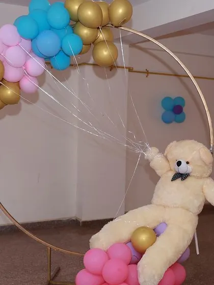 A creative baby shower decoration featuring a large teddy bear appearing to swing from a circular frame decorated with balloons.