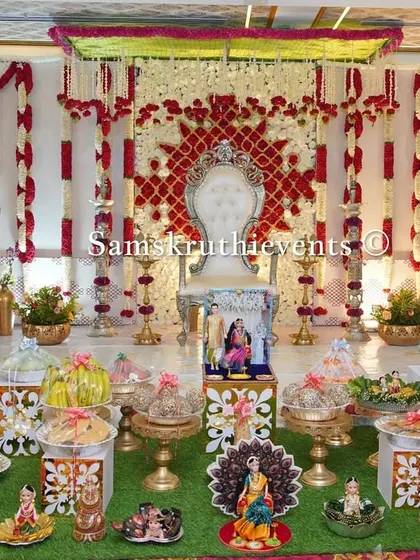 A beautiful Seemantha setup with a red and white floral backdrop. The artificial grass flooring and detailed doll arrangements in the foreground add a touch of charm and tradition to the event.